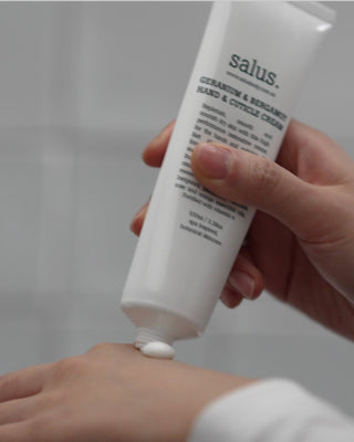 Hand applying cream from a 'salus' tube to a wrist against a neutral background