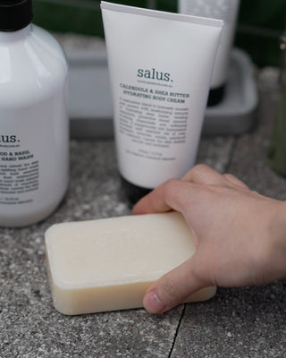 Hand holding a bar of soap with 'salus' body cream and bottle in the background.