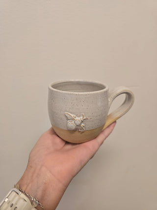 Hand holding a ceramic mug with a bee design against a plain background