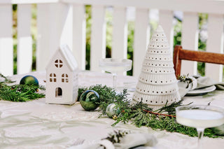 Decorative table setting with white ceramic house and tree, green ornaments, and evergreen branches on a white tablecloth.