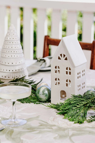 Decorative table setting with white ceramic house-shaped lantern, greenery, and glasses on a white tablecloth.