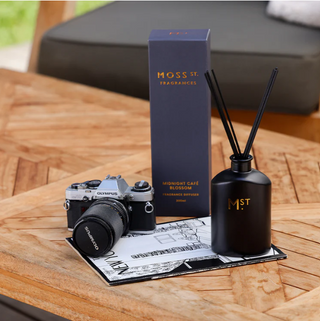 Moss St. diffuser and packaging on a wooden table with a camera and newspaper.