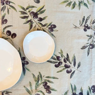 Table setting with white bowls on a tablecloth featuring olive branches and olives.