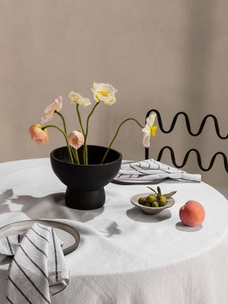 Table setting with a black bowl of flowers, a small bowl of olives, and a peach on a white tablecloth.