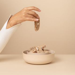 Hand holding a piece of bread above a bowl filled with bread pieces on a beige background