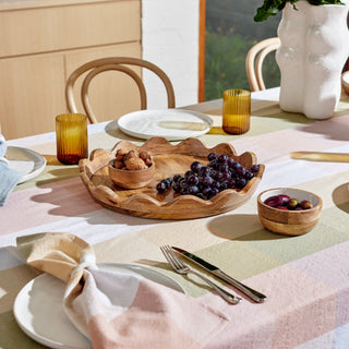Dining table setting with a wooden bowl of grapes and other fruits on a colorful tablecloth.