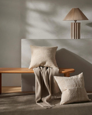 Beige pillows and a blanket on a wooden bench with a lamp in the background
