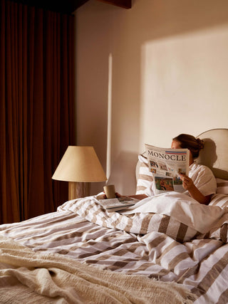Person reading a newspaper in bed with a lamp and curtains in the background