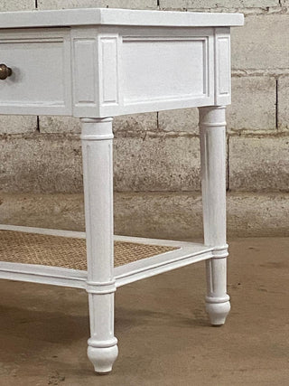 White wooden side table with a woven shelf against a stone wall.