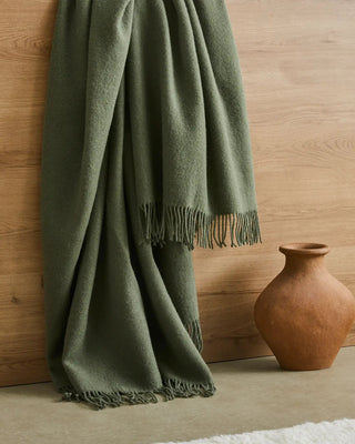 Green blanket with fringes draped over a wooden surface next to a terracotta vase.