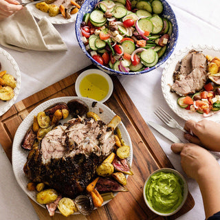 Dinner table with roasted meat, vegetables, and salads on a wooden cutting board.