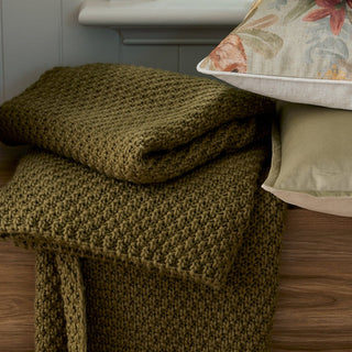Folded green blanket on a wooden surface with a floral pillow in the background