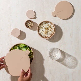Beige salad bowls with lids on a light surface, containing salad and other ingredients.