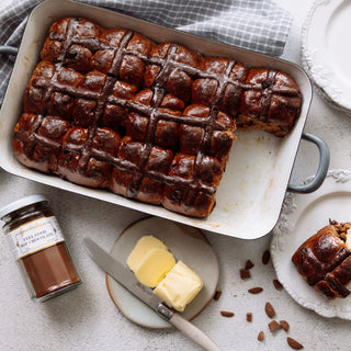 Hot cross buns in a baking dish with a jar of chocolate spread and butter on a table.