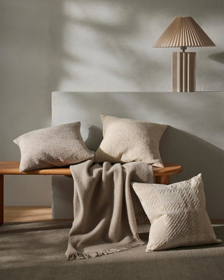 Beige pillows and a blanket on a wooden bench with a lamp in the background