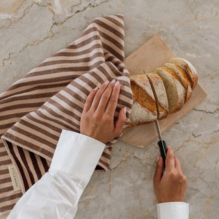 Person slicing bread on a wooden board with a striped towel nearby on a marble countertop.
