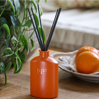 Orange diffuser with black sticks on a wooden surface with a plant and oranges in the background