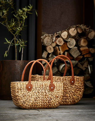 Two woven bags with leather handles on a wooden surface with a stack of firewood in the background.