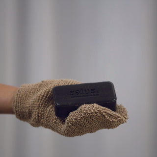 Hand holding a bar of black soap with 'salus' branding on a beige textured cloth against a neutral background.