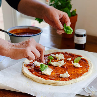 Person adding basil leaves to a pizza with tomato sauce and cheese on a wooden table.