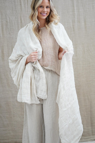Woman wearing a white shawl over a beige sweater and pants against a neutral background