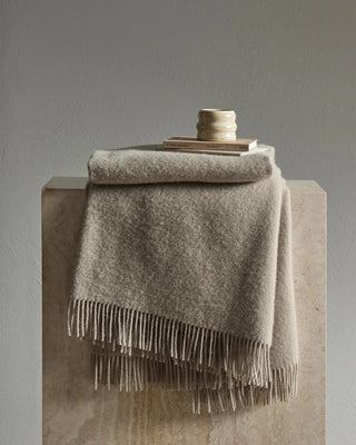 Gray throw blanket with fringe draped over a wooden surface against a gray background