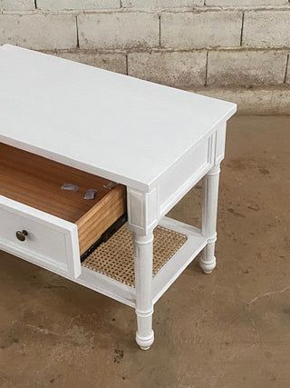 White wooden table with a drawer and wicker shelf against a brick wall.