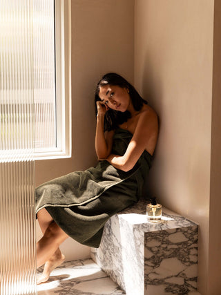 Woman wrapped in a towel sitting by a window in a softly lit room.