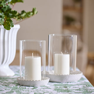 Two white candles in clear glass holders on a decorative white base and decorative tablecloth with a blurred background