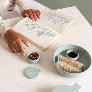 Person reading a book with a bowl of snacks and a small container on a light surface.