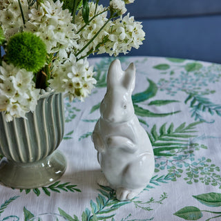 White ceramic rabbit figurine next to a vase with white flowers on a tablecloth with green leaf pattern