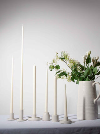 Set of white candles with decorative holders on a light surface, with a vase of flowers in the background.