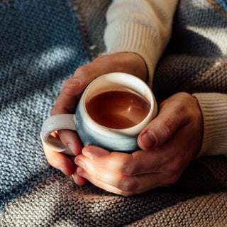 Person holding a mug of hot chocolate with a cozy blanket in the background