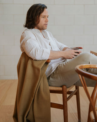 Man sitting on a wooden chair holding a glass, with a beige blanket draped over his lap.