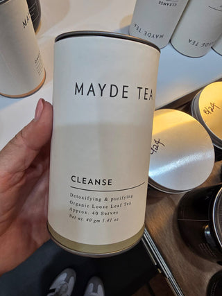 Mayde Tea Cleanse canister held by a hand with more tea containers in the background.