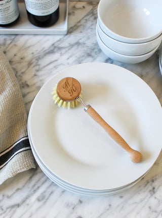 Dishwashing brush with wooden handle and monogram on a white plate, with stacked bowls and a towel in the background.