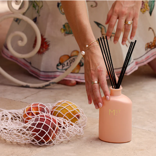 Pink diffuser with black reeds being held by a hand, with fruits in the background.