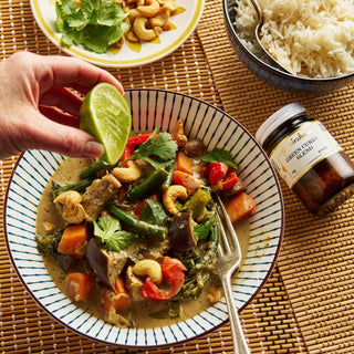 Person squeezing lime over a bowl of curry with vegetables and cashews, on a woven mat with a bottle of curry paste.