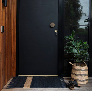 Black door with a doormat, plant, and shoes on a wooden floor.