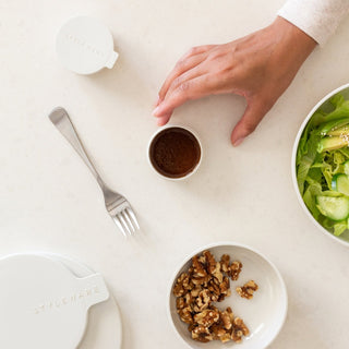 Hand reaching for a small container of dressing on a table with a salad and nuts.