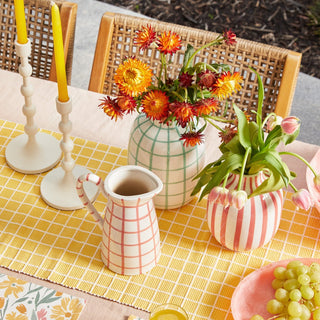 Table setting with flowers, candles, and a pitcher on a yellow checkered tablecloth.