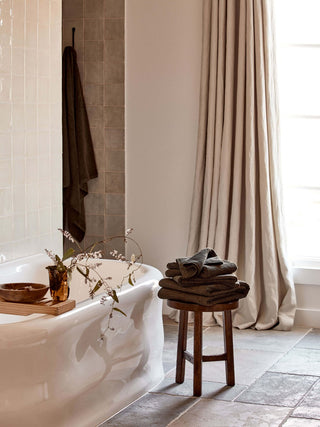 Bathroom with a white bathtub, wooden stool with towels, and large window with curtain.