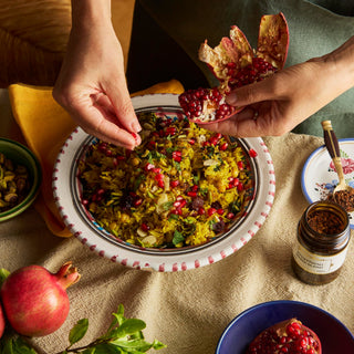 Person adding pomegranate seeds to a dish of rice on a table with pomegranates and a jar.