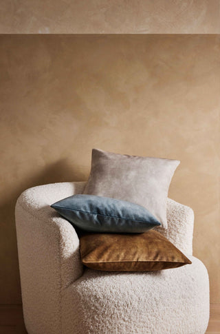 White armchair with three pillows in gray, blue, and brown against a beige wall.