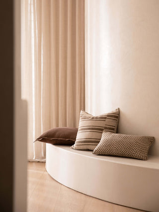 Three pillows on a curved white bench against a neutral wall with curtains.