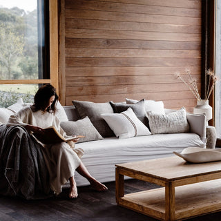 Woman reading a book on a gray sofa in a modern living room with wooden walls and a coffee table.