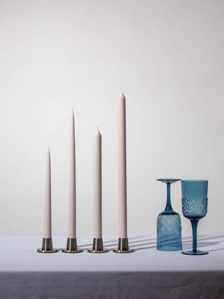 Set of four tall candles with metallic holders on a white surface, accompanied by two blue glass goblets.