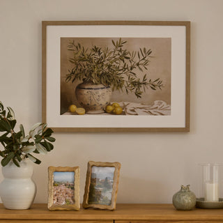 Framed artwork of a still life with a vase and lemons on a wall above a wooden shelf with decorative items.