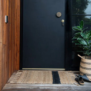 Black door with a doormat, plant, and shoes on a wooden porch.
