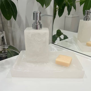 Soap dispenser with a bar of soap on a white surface, with a mirror and plant in the background.